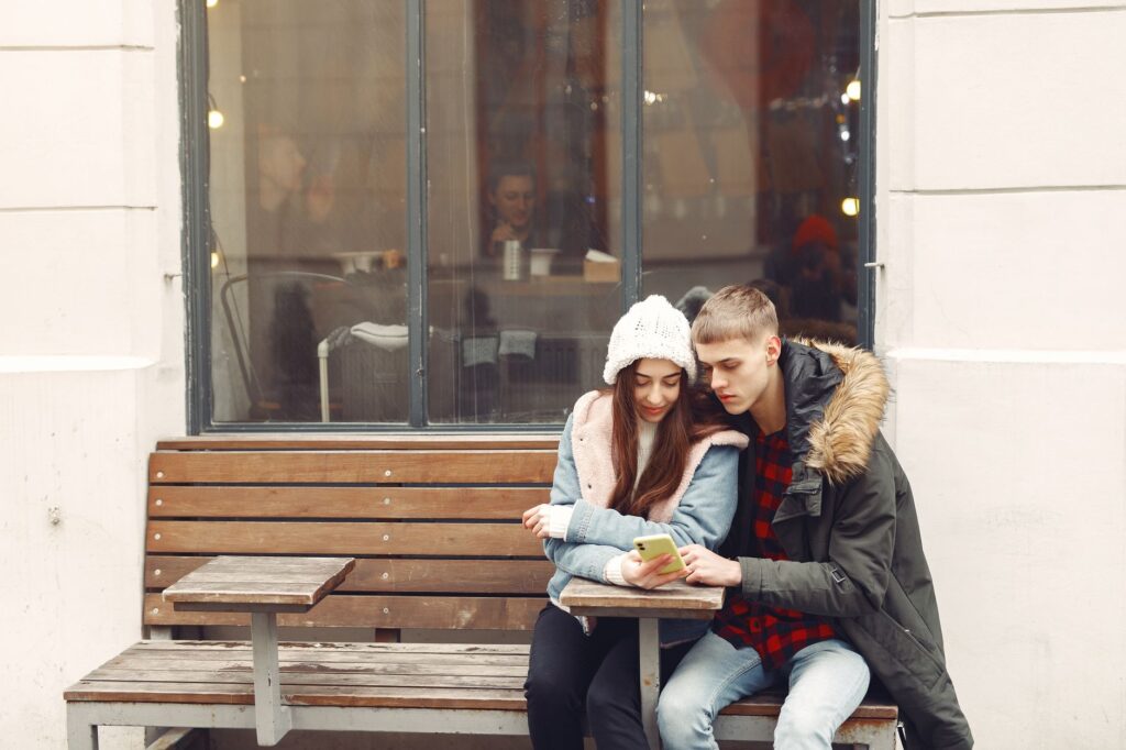 A young couple in warm clothing looks at a smartphone together on a bench outside a cafe.